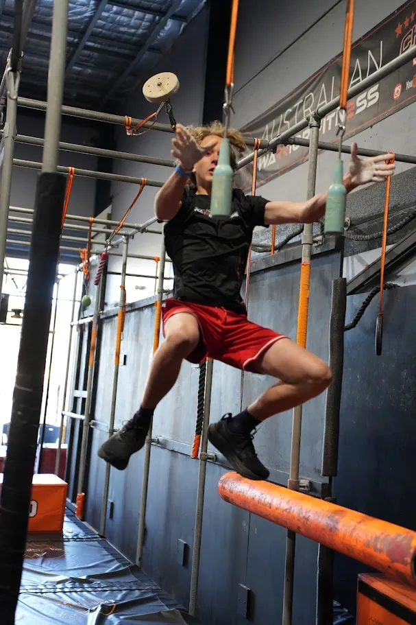 Competitor in black and red swinging from grips above a log obstacle