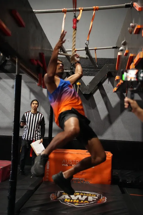 Competitor in orange swinging on the rig as a referee watches